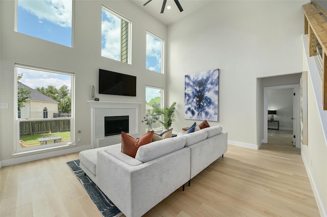 living area featuring a fireplace with raised hearth, ceiling fan, light wood-style flooring, a towering ceiling, and baseboards