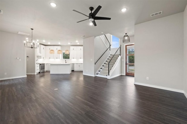 unfurnished living room with ceiling fan with notable chandelier, dark wood-type flooring, visible vents, baseboards, and stairs