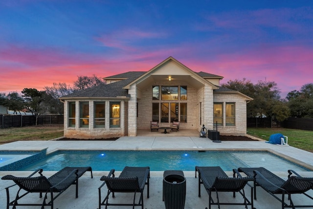 back of house at dusk with stone siding, a pool with connected hot tub, a patio area, and fence