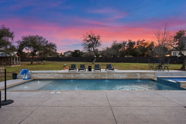 pool at dusk with a yard, a patio, fence, and a pool with connected hot tub