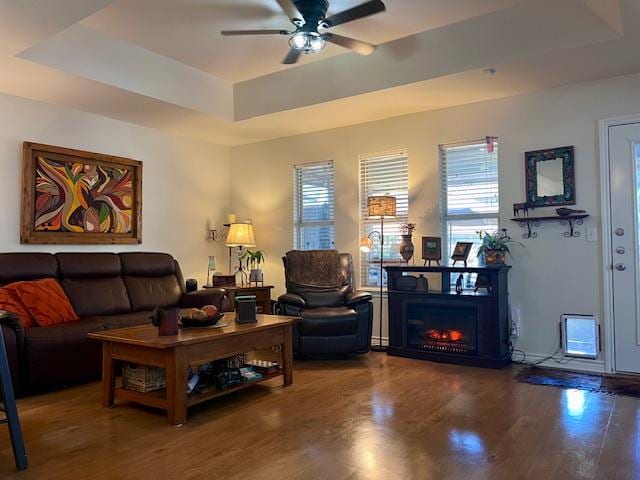 living area with a tray ceiling, ceiling fan, a lit fireplace, and wood finished floors