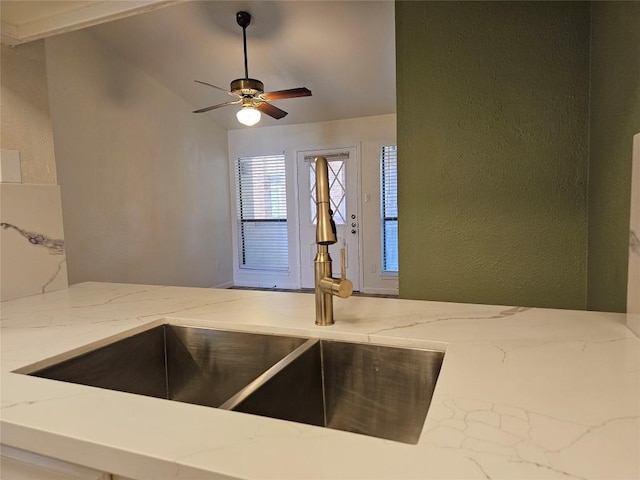 kitchen featuring lofted ceiling, a textured wall, light stone counters, ceiling fan, and a sink