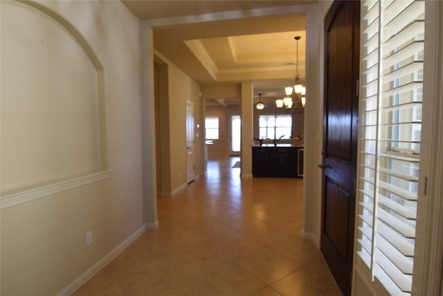 corridor featuring baseboards, a tray ceiling, light tile patterned flooring, and an inviting chandelier