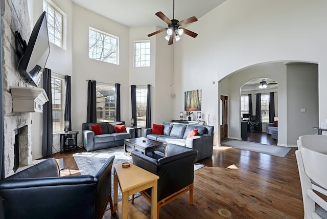 living room featuring arched walkways, a ceiling fan, a stone fireplace, wood finished floors, and baseboards