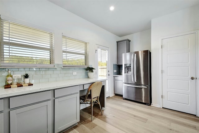 kitchen featuring gray cabinets, backsplash, stainless steel refrigerator with ice dispenser, light stone counters, and light wood-style flooring