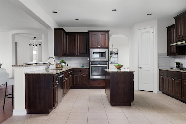 kitchen featuring light stone counters, appliances with stainless steel finishes, dark brown cabinetry, a sink, and a kitchen island