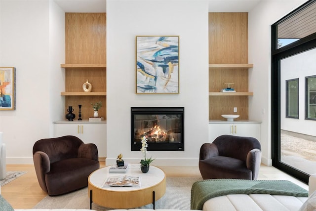 sitting room with a glass covered fireplace, built in shelves, light wood-type flooring, and baseboards