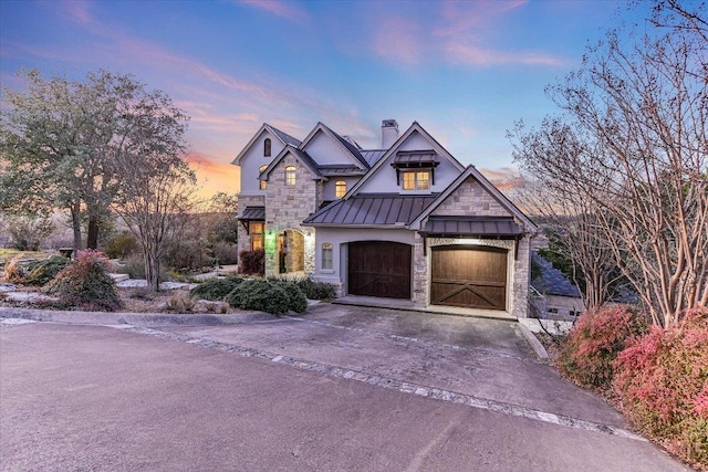 view of front facade with metal roof, driveway, stone siding, a standing seam roof, and a chimney