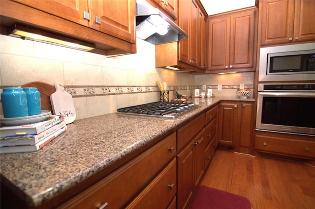 kitchen with dark wood-style floors, stainless steel appliances, tasteful backsplash, brown cabinetry, and under cabinet range hood