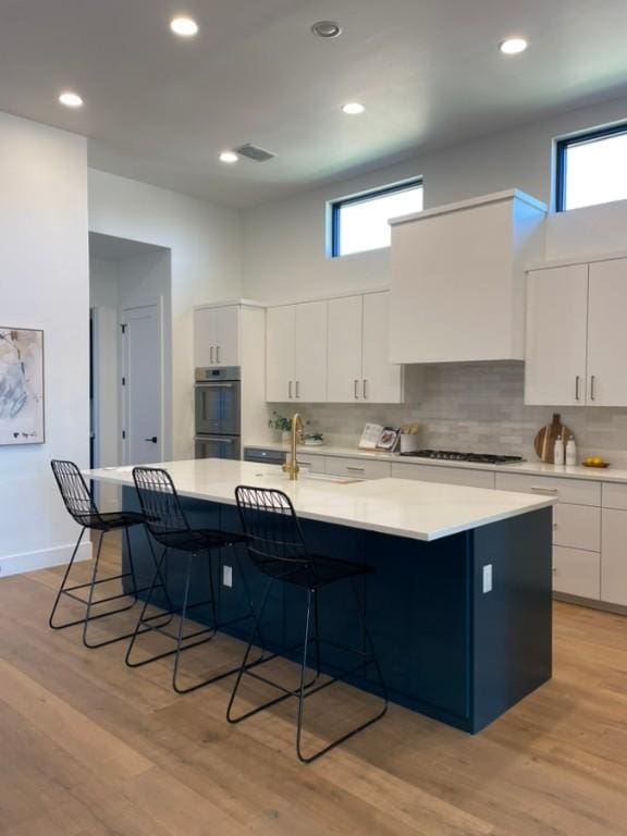 kitchen with double oven, light countertops, light wood finished floors, a kitchen bar, and a large island with sink