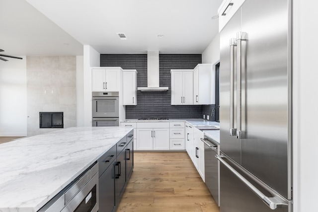 kitchen with light stone counters, stainless steel appliances, white cabinetry, visible vents, and wall chimney range hood