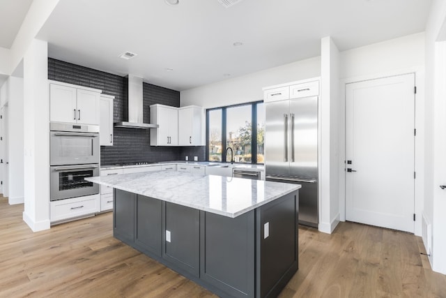 kitchen featuring white cabinets, wall chimney exhaust hood, a kitchen island, appliances with stainless steel finishes, and light wood-type flooring