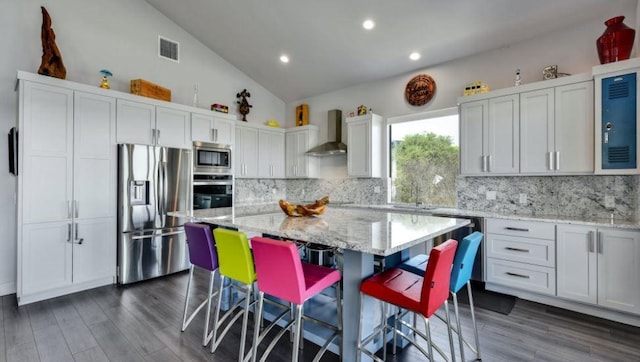 kitchen featuring wall chimney exhaust hood, light stone counters, appliances with stainless steel finishes, vaulted ceiling, and a sink