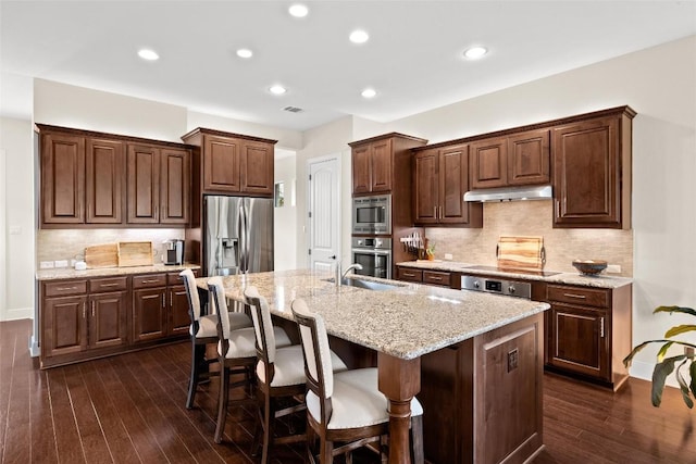 kitchen with an island with sink, appliances with stainless steel finishes, dark wood-style flooring, under cabinet range hood, and a sink