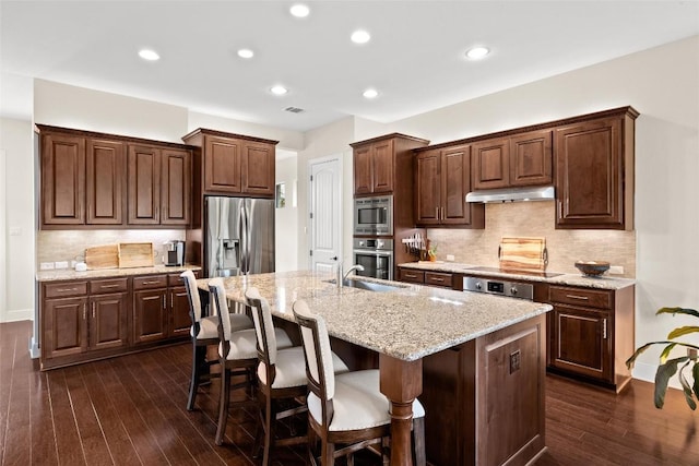 kitchen featuring decorative backsplash, a kitchen breakfast bar, appliances with stainless steel finishes, a center island with sink, and under cabinet range hood