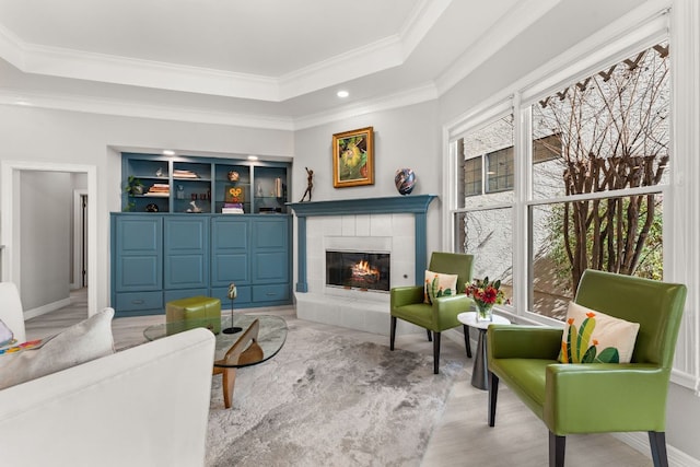 living area featuring light wood-style flooring, baseboards, ornamental molding, a tray ceiling, and a tiled fireplace