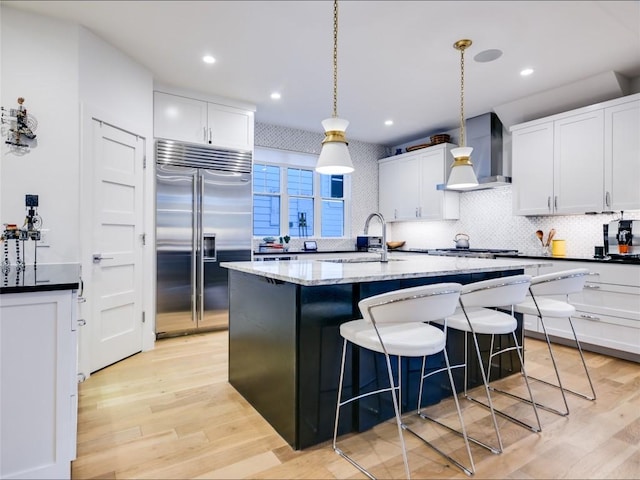 kitchen with stainless steel appliances, a breakfast bar, a sink, wall chimney range hood, and a center island with sink