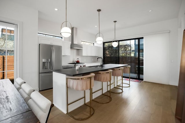 kitchen featuring wood finished floors, a sink, white cabinetry, wall chimney range hood, and stainless steel refrigerator with ice dispenser