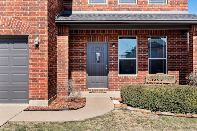 view of exterior entry featuring an attached garage, brick siding, and roof with shingles