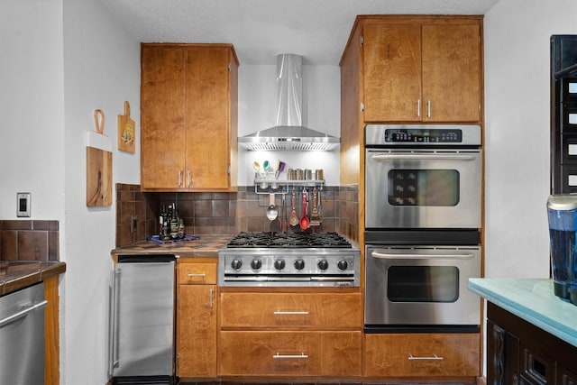 kitchen featuring tile counters, decorative backsplash, wall chimney exhaust hood, appliances with stainless steel finishes, and brown cabinets