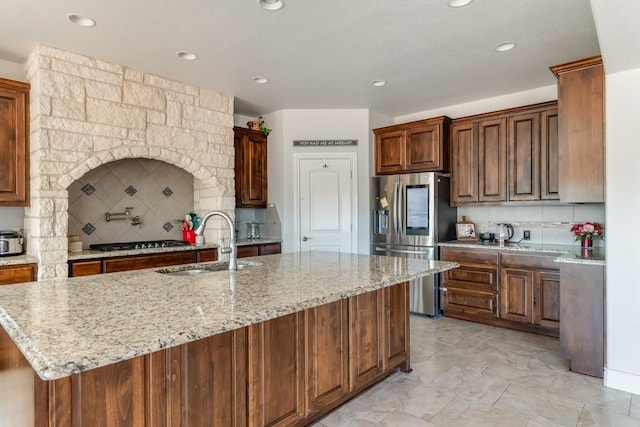 kitchen with a sink, gas stovetop, stainless steel refrigerator with ice dispenser, decorative backsplash, and a center island with sink