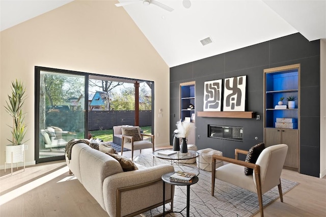 living room with high vaulted ceiling, built in shelves, visible vents, light wood-type flooring, and a glass covered fireplace