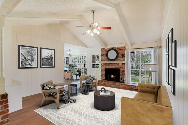 living area with a ceiling fan, lofted ceiling with beams, wood finished floors, a textured ceiling, and a brick fireplace