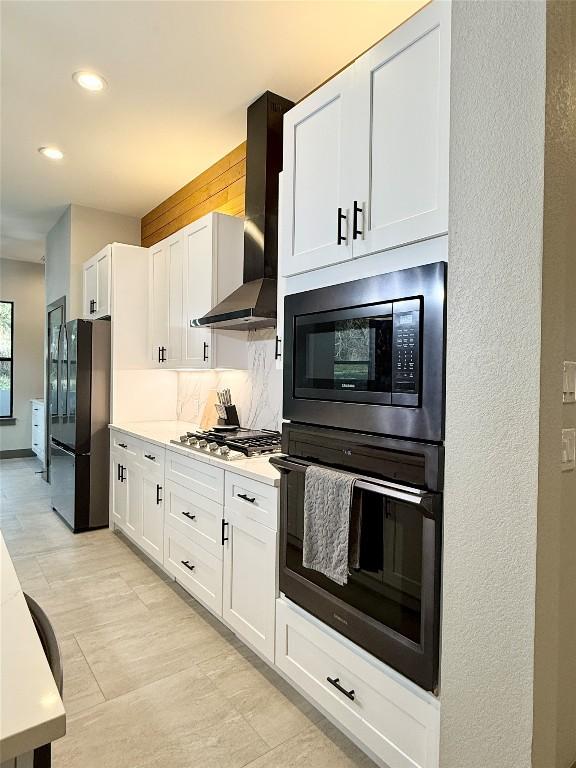 kitchen featuring white cabinets, wall chimney range hood, light countertops, black appliances, and backsplash