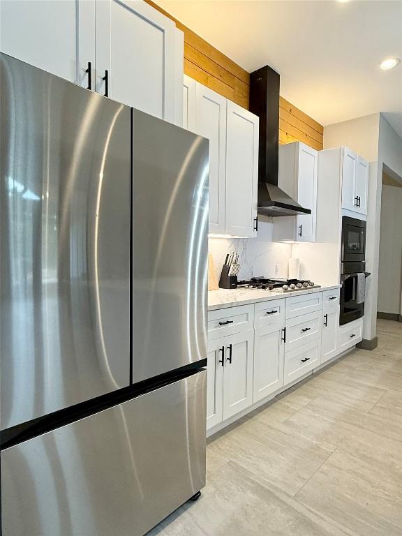 kitchen featuring light stone counters, wall chimney range hood, black appliances, white cabinetry, and backsplash