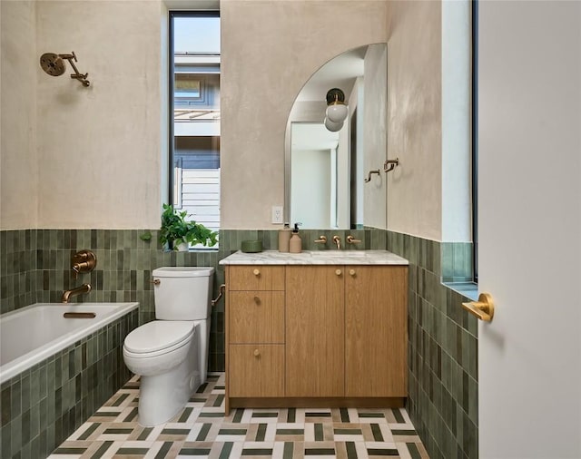 bathroom with a wainscoted wall, tile walls, toilet, vanity, and tiled tub