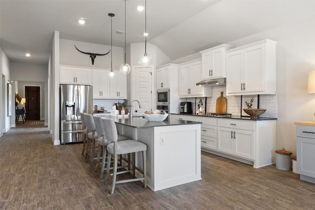 kitchen with dark wood-style flooring, stainless steel appliances, dark countertops, under cabinet range hood, and a kitchen bar