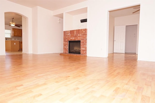 unfurnished living room featuring light wood finished floors, ceiling fan, a brick fireplace, and arched walkways