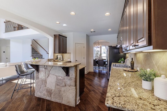 kitchen featuring arched walkways, light stone counters, a kitchen breakfast bar, backsplash, and dark wood-style floors