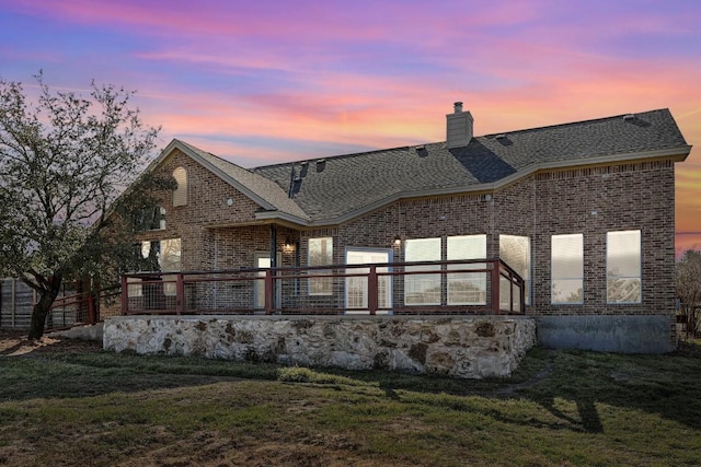 back of house at dusk with brick siding, a yard, a chimney, and roof with shingles