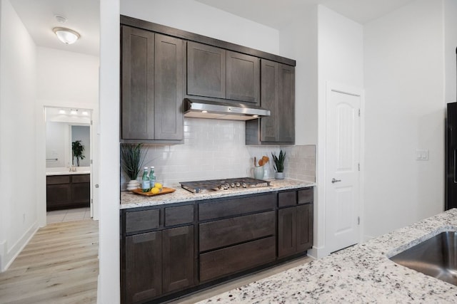 kitchen featuring light stone counters, dark brown cabinets, under cabinet range hood, stainless steel gas cooktop, and backsplash