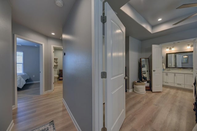 corridor featuring a tray ceiling, light wood finished floors, recessed lighting, a sink, and baseboards