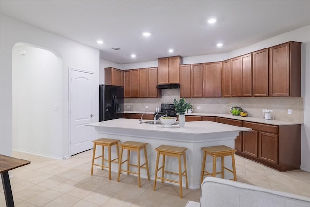 kitchen featuring arched walkways, black fridge with ice dispenser, decorative backsplash, under cabinet range hood, and a kitchen bar