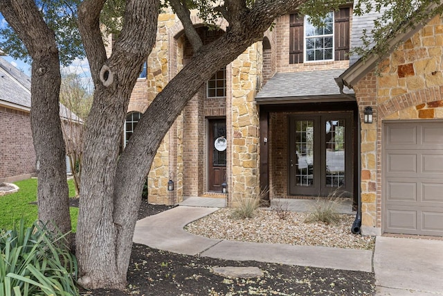 doorway to property featuring french doors, roof with shingles, brick siding, an attached garage, and stone siding