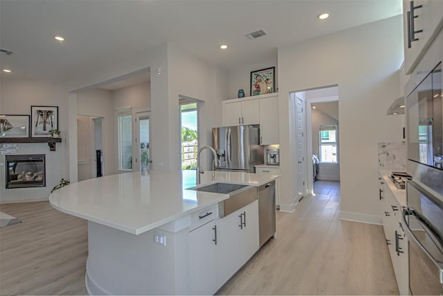 kitchen with light wood finished floors, appliances with stainless steel finishes, a glass covered fireplace, white cabinetry, and a sink