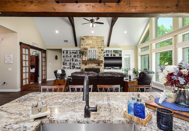 kitchen featuring a ceiling fan, wood finished floors, beamed ceiling, a stone fireplace, and high vaulted ceiling