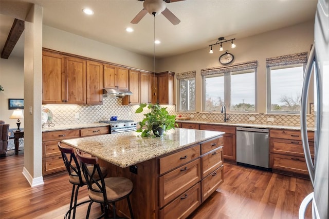 kitchen featuring dark wood finished floors, brown cabinetry, stainless steel appliances, under cabinet range hood, and a sink