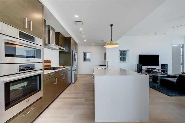 kitchen with wall chimney range hood, modern cabinets, appliances with stainless steel finishes, and a kitchen island with sink