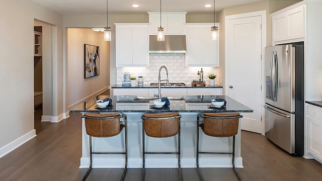 kitchen featuring stainless steel fridge with ice dispenser, dark wood-style floors, a breakfast bar, a kitchen island with sink, and under cabinet range hood