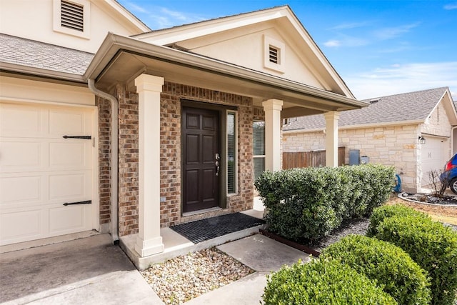 entrance to property featuring an attached garage, brick siding, roof with shingles, and stucco siding