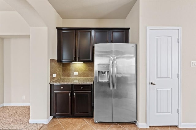 kitchen with dark brown cabinetry, arched walkways, decorative backsplash, light stone counters, and stainless steel refrigerator with ice dispenser