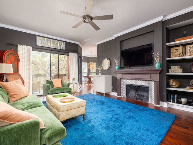 living room with built in shelves, ornamental molding, a ceiling fan, wood finished floors, and a tile fireplace