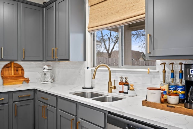 kitchen with tasteful backsplash, stainless steel dishwasher, gray cabinetry, a sink, and light stone countertops