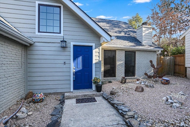 property entrance with roof with shingles, a chimney, fence, and brick siding