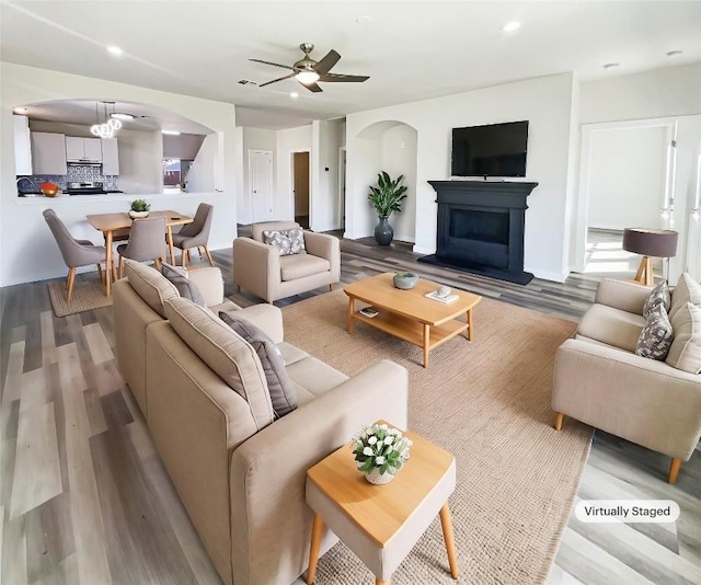 living area featuring arched walkways, a fireplace with raised hearth, ceiling fan, and wood finished floors