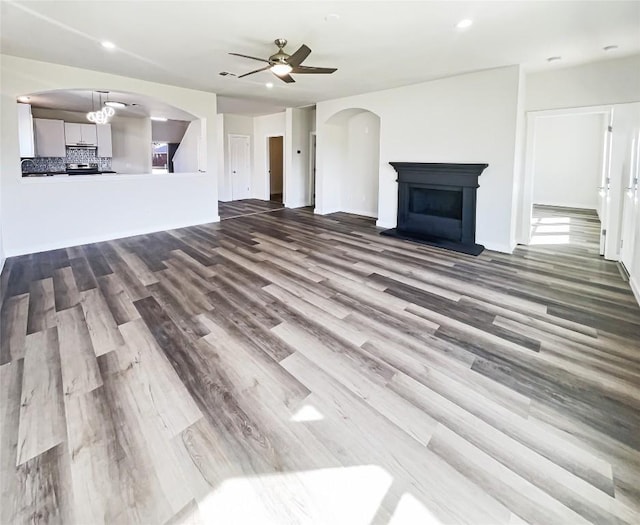 unfurnished living room with arched walkways, a fireplace with raised hearth, ceiling fan, and wood finished floors